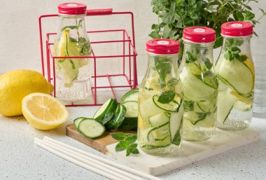 Kırmızı kapaklı cam şişeler, içinde salatalık, limon dilimleri ve nane yaprakları bulunan detoks suyla dolu beyaz masa.