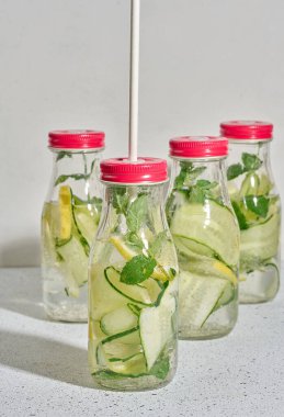 Kırmızı kapaklı cam şişeler, içinde salatalık, limon dilimleri ve nane yaprakları bulunan detoks suyla dolu beyaz masa.