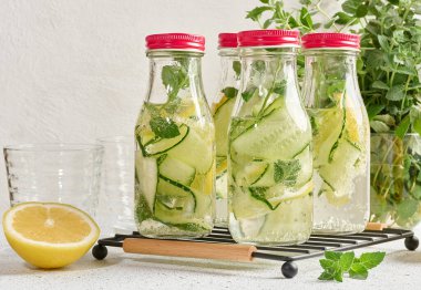 Kırmızı kapaklı cam şişeler, içinde salatalık, limon dilimleri ve nane yaprakları bulunan detoks suyla dolu beyaz masa.