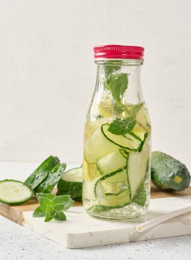 Kırmızı kapaklı cam şişeler, içinde salatalık, limon dilimleri ve nane yaprakları bulunan detoks suyla dolu beyaz masa.