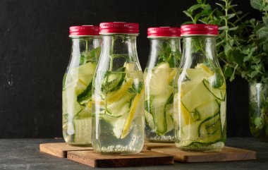 İçi ferahlatıcı su dolu kırmızı kapaklı cam şişeler, içinde salatalık ve limon dilimleri ve tahta bir tahtanın üzerinde nane yaprakları var.