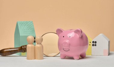 Pembe kumbara, ahşap evler, tahta insan figürleri ve büyüteç. Gayrimenkul, konut yatırımları veya aile bütçesi finans planlaması