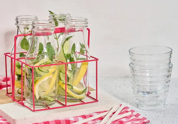 Salatalık, limon dilimleri ve nane yaprakları içeren detoks suyuyla dolu cam şişeler kesme tahtasındaki kırmızı metal bir tutacağa konmuş. Doldurulmuş boş bardaklar ve metal pipetler de görünür