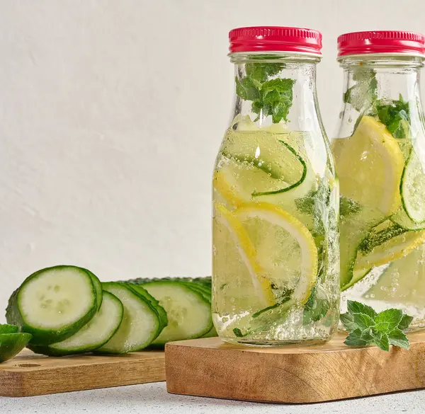 Kırmızı kapaklı cam şişeler, içinde salatalık, limon dilimleri ve nane yaprakları bulunan detoks suyla dolu beyaz masa.