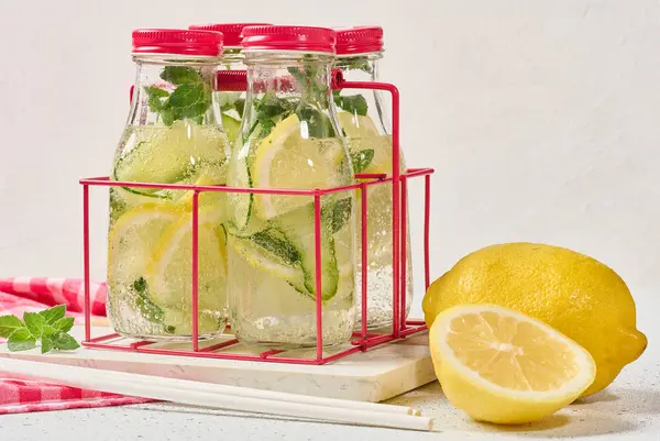 Kırmızı kapaklı cam şişeler, içinde salatalık, limon dilimleri ve nane yaprakları bulunan detoks suyla dolu beyaz masa.