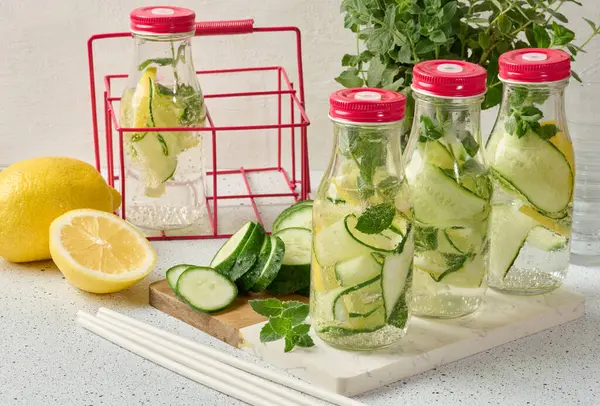 Kırmızı kapaklı cam şişeler, içinde salatalık, limon dilimleri ve nane yaprakları bulunan detoks suyla dolu beyaz masa.