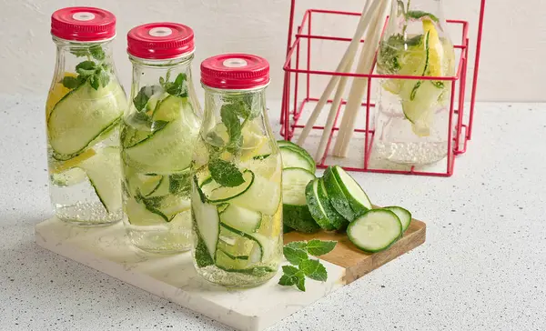 Kırmızı kapaklı cam şişeler, içinde salatalık, limon dilimleri ve nane yaprakları bulunan detoks suyla dolu beyaz masa.