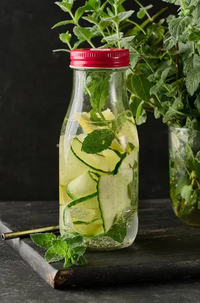 İçi salatalık, limon dilimleri ve nane yapraklarıyla dolu cam şişe. Şişe koyu bir tahtanın üzerinde.