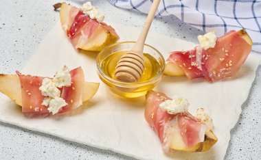 Beyaz bir tahtada peynirli jambona sarılı armut dilimi ve susam tohumları.