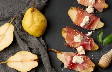 Bir dilim armut jambona sarılmış, üstü peynir ve susam tohumlarıyla kaplı, siyah bir levhada, üst görünümde.