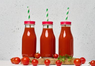 Kırmızı başlıklı domates suyu ile dolu cam şişeler. Taze kırmızı domatesler ahşap bir standın üzerinde ve önlerinde beyaz bir zemin var.