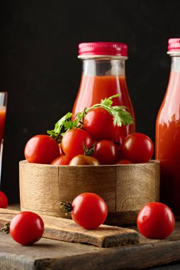 Tahta bir kasede kırmızı kiraz domatesleri. Domatesler ahşap bir tahtaya saçılmış, domates suyu şişeleri yakınlarda.