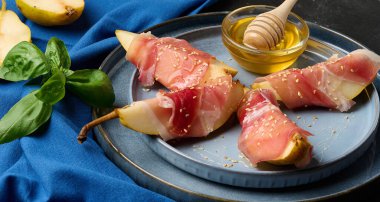 Bir armut aperatifi Prosciutto, susam tohumları serpiştirilmiş ve altın bal ile servis edilmiş. Tabak mavi bir tabakta taze fesleğen yapraklarının yanında servis edilir..