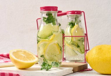 Kırmızı kapaklı cam şişeler, içinde salatalık, limon dilimleri ve nane yaprakları bulunan detoks suyla dolu beyaz masa.