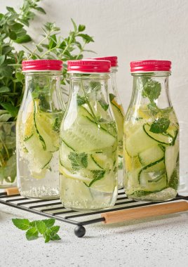 Kırmızı kapaklı cam şişeler, içinde salatalık, limon dilimleri ve nane yaprakları bulunan detoks suyla dolu beyaz masa.
