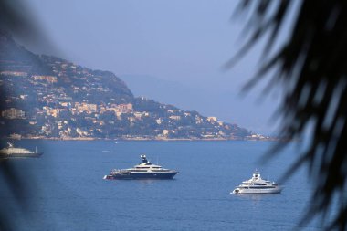 Saint-Jean-Cap-Ferrat, Fransa - 29 Temmuz 2021: Saint-Jean-Cap-Ferrat yarımadasından Akdeniz 'e ve gemilere, ön planda palmiye yaprakları odak dışı