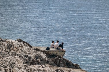 Saint-Jean-Cap-Ferrat, Fransa - 01 Haziran 2023: Kayalık bir kıyıda oturup denize bakan üç kişi