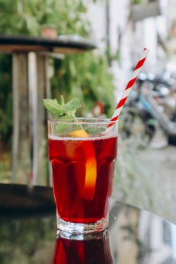 Bir bardak kırmızı içecek (meyveli buzlu çay) ve bir pipet, şehirdeki bir kafenin terasında.