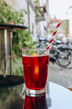 Bir bardak kırmızı içecek (meyveli buzlu çay) ve bir pipet, şehirdeki bir kafenin terasında.
