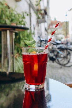 Bir bardak kırmızı içecek (meyveli buzlu çay) ve bir pipet, şehirdeki bir kafenin terasında.