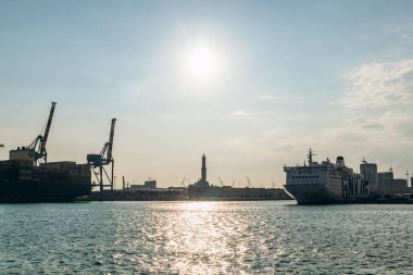 Cenova, İtalya - 22 Eylül 2024: Lanterna deniz fenerinin silueti, konteynır vinçleri ve gün batımında işlek limandaki yolcu gemisi.