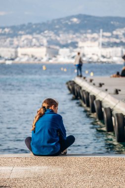 Cannes, Fransa - 8 Mart 2025: Sainte-Marguerite Adası 'nda deniz kenarında oturan ve arkasında Cannes şehri ve dağları olan kişi.
