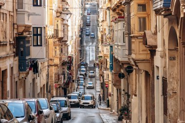 Valletta, Malta - 9 Nisan 2025: Kireçtaşı binalar ve renkli Malta balkonları, park edilmiş arabalar ve bahar şehri hayatı ile kaplı güneşli dar bir cadde