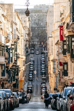 Valletta, Malta - 9 Nisan 2025: Kireçtaşı binalar ve renkli Malta balkonları, park edilmiş arabalar ve bahar şehri hayatı ile kaplı güneşli dar bir cadde