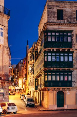 Valletta, Malta - 9 Nisan 2025: Parlayan ışıklar, geleneksel Malta balkonları, tarihi kireçtaşı binalar, park edilmiş arabalar ve tepeye tırmanan insanlarla dolu akşam caddesi