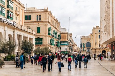Valletta, Malta - 10 Nisan 2025: Zarif kireçtaşı binalar ve geleneksel yeşil balkonlarla çevrili şehir meydanı, ağaçların ve modern parlamento cephesinin önünden geçen kalabalıklar