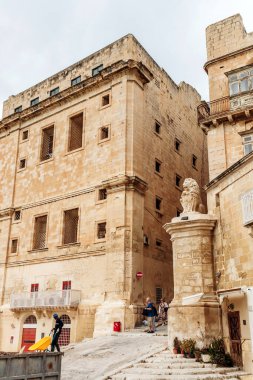 Valletta, Malta - 10 Nisan 2025: Demirden pencereli güçlendirilmiş kireçtaşı bina, yayalar tarihi yamaçtan geçerken bir aslanın tepesinde duruyor.