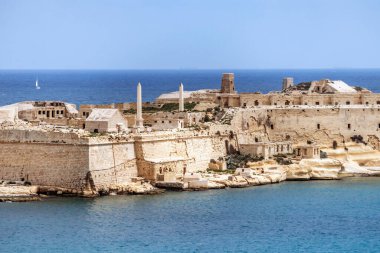 Saint Elmo kıyı tahkimatı, kireçtaşı surları ve Akdeniz kıyısında tarihi deniz duvarlarının yakın çekim görüntüsü. Arka planda mavi su ve yelkenli tekneler var..