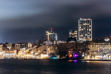 St Julians, Malta - 10 Nisan 2025: Modern kıyı şeridi geceleri ışıldayan gökdelenler, parlayan oteller, Balluta Körfezi kıyısı boyunca palmiye ağaçları