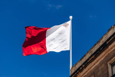 Malta 'nın ulusal bayrağı, üst köşesinde George Cross olan kırmızı ve beyaz, açık mavi gökyüzünde geleneksel Malta kireçtaşı mimarisinin üzerinde dalgalanır.