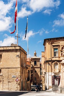 Birgu, Malta - 11 Nisan 2025: Bal rengi kireç taşı binalar, heykel süsü, St John Şövalyeleri de dahil olmak üzere dalgalanan bayraklar ve tarihi balkonlarla çevrili güneşli kasaba meydanı