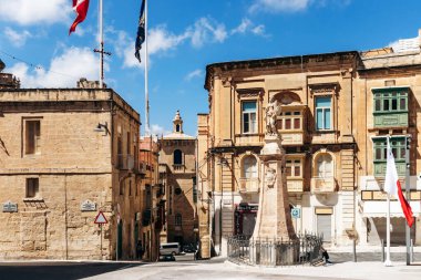 Birgu, Malta - 11 Nisan 2025: Bal rengi kireç taşı binalar, heykel süsü, St John Şövalyeleri de dahil olmak üzere dalgalanan bayraklar ve tarihi balkonlarla çevrili güneşli kasaba meydanı