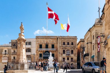 Birgu, Malta - 11 Nisan 2025: İnsanlarla dolu parlak kasaba meydanı, tarihi kireçtaşı cepheleri, iki önemli heykel, ve Aziz John Şövalyeleri, Vatikan ve Malta Şovalyeleri bayrakları