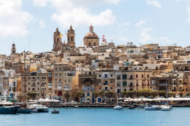Geleneksel kireçtaşı binaları, renkli tekneleri ve deniz kenarındaki kafeleri parlak bir gökyüzü ve büyük kilise kubbeleri altında yer alan resimli Malta limanı.