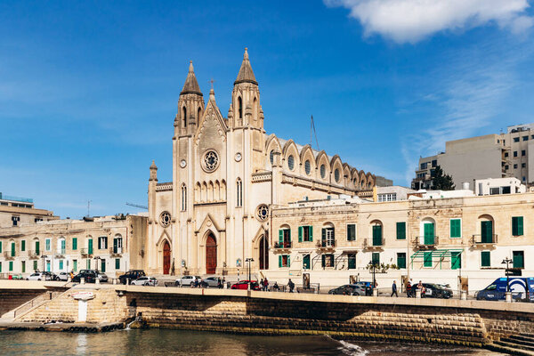 Баллута-Бей, Мальта - 9 апреля 2025 года: Неоготическая кармелитовская церковь стоит на набережной залива Баллута между заливом Святого Джулиана и Слимой, окруженная традиционными и современными зданиями