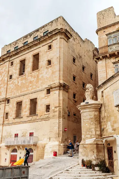 Valletta, Malta - 10 Nisan 2025: Demirden pencereli güçlendirilmiş kireçtaşı bina, yayalar tarihi yamaçtan geçerken bir aslanın tepesinde duruyor.