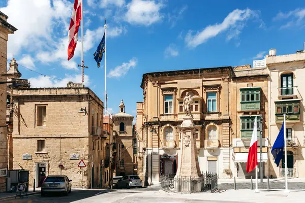 Birgu, Malta - 11 Nisan 2025: Tarihi kireçtaşı binalar, merkezi taş heykel, Malta, Avrupa 'nın canlı bayrakları ve St. John Şövalyeleri tarafından çerçevelenmiş güneşli meydan