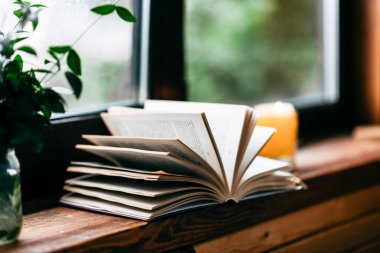 Açık bir kitap pencerenin kenarında duruyor, pencerenin dışında yağmur damlaları, sarı bir mum yanıyor, vazoda yeşil bir buket, rahat bir atmosfer, sonbahar havası, okuma zamanı.