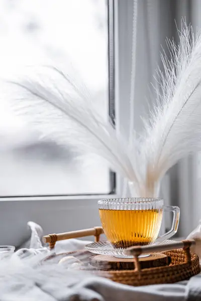 Şeffaf bir fincanda altın siyah çay. Hasır bir tepside bir fincan çay. Beyaz bir pencere kenarında, hafif bir kompozisyon, altın ambalajlı şekerler.