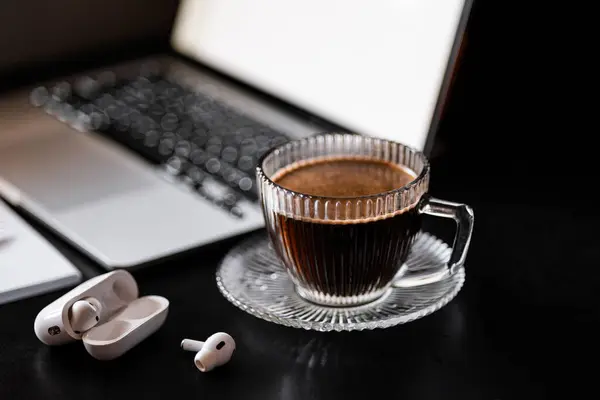 transparent cup with steaming black coffee on a black desktop near a laptop and white headphones, business, receiving clients in the office, notebook, work environment, open laptop in the background