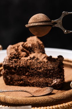Bir parça kremalı çikolatalı pasta üzerine metal bir süzgeçten kakao serpiştiriyor. Tahta tabakta çikolatalı pasta ve beyaz kahve masasında hasır tepsi, koyu arkaplan, doğum günü pastası.