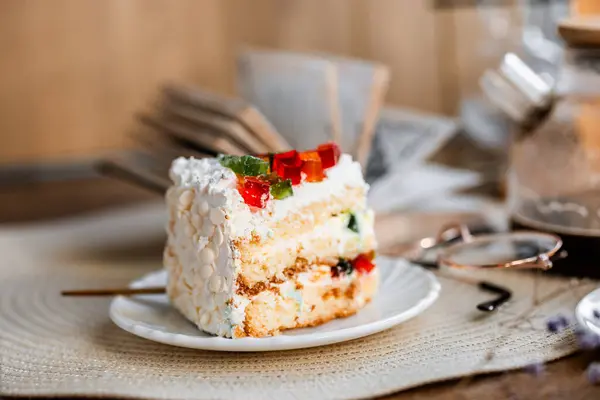 a piece of cake with buttercream on pieces of multi-colored jelly on a white saucer on a wooden tray under the sun's rays, espresso coffee in a white cup, a transparent teapot with coffee, flowers