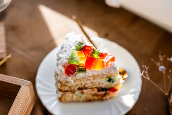 a piece of cake with buttercream on pieces of multi-colored jelly on a white saucer on a wooden tray under the sun's rays, espresso coffee in a white cup, a transparent teapot with coffee, flowers