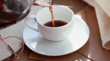Espresso kahvesi beyaz bir fincana konur. Güneş ışınlarının altındaki ahşap bir tepside, şeffaf bir çaydanlık kahve, çiçekler, bir kitap, bir sabah kahvesi, bir içecek, hafif bir kompozisyon.