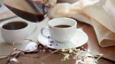 Espresso kahvesi beyaz bir fincana konur. Güneş ışınlarının altındaki ahşap bir tepside, şeffaf bir çaydanlık kahve, çiçekler, bir kitap, bir sabah kahvesi, bir içecek, hafif bir kompozisyon.