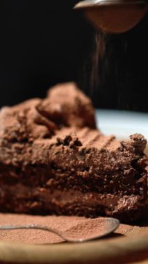 Bir parça kremalı çikolatalı pasta üzerine metal bir süzgeçten kakao serpiştiriyor. Tahta tabakta çikolatalı pasta ve beyaz kahve masasında hasır tepsi, koyu arkaplan, doğum günü pastası.